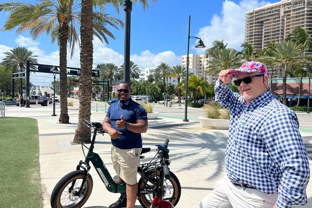 Two men one giving a thumbs up and the other holding his cap are standing beside their bicycles on a sunny day with palm trees and urban structures in the background