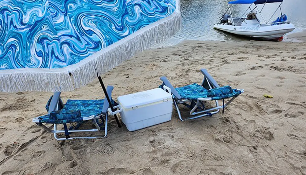 Two blue beach chairs and a cooler are placed on the sand in front of a boat with a tie-dye patterned towel hanging above to provide shade