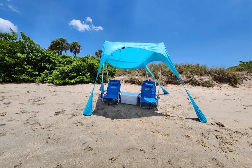 A blue beach canopy shades two chairs and a cooler on a sandy beach with lush greenery in the background under a clear blue sky