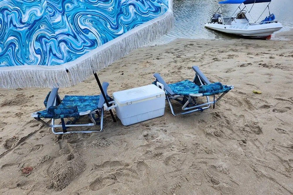Two beach chairs with blue cushions and a cooler positioned on sandy shore near a boat with a patterned beach blanket hanging in the background