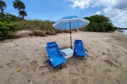 A relaxing beach setup featuring two blue lounge chairs and a cooler under a patterned umbrella on sandy shore.