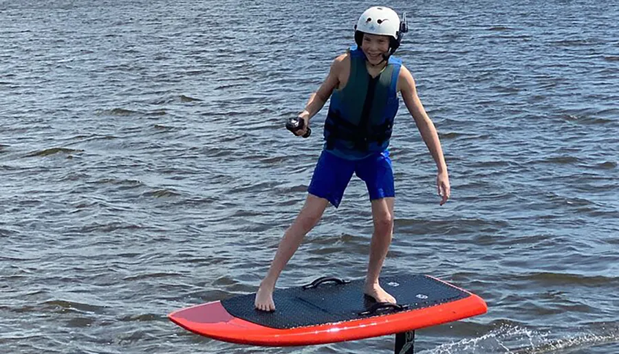 A child wearing a helmet and life vest is smiling while balancing on a hydrofoil surfboard above the water.