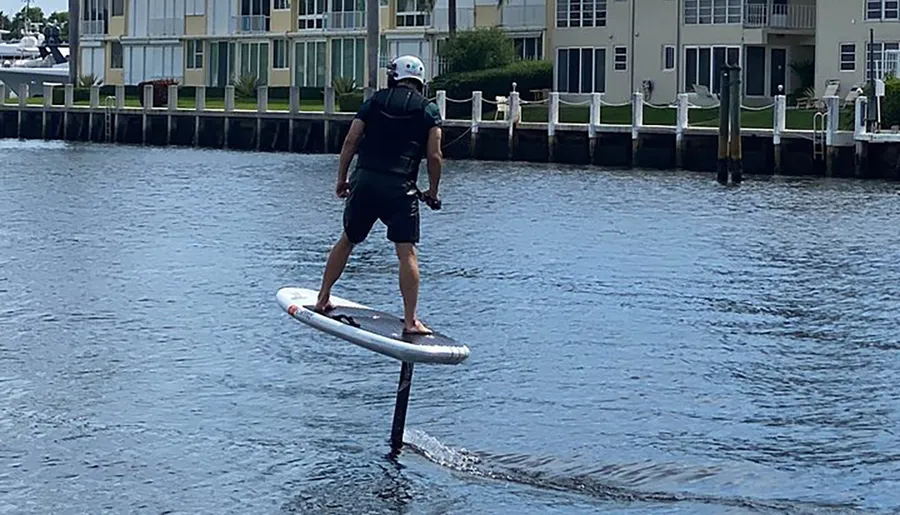 A person is hydrofoiling on a body of water against a backdrop of residential buildings.
