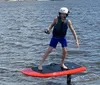A smiling person is standing on a motorized surfboard above the water wearing a helmet and life vest