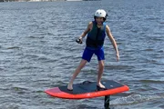 A smiling person is standing on a motorized surfboard above the water, wearing a helmet and life vest.