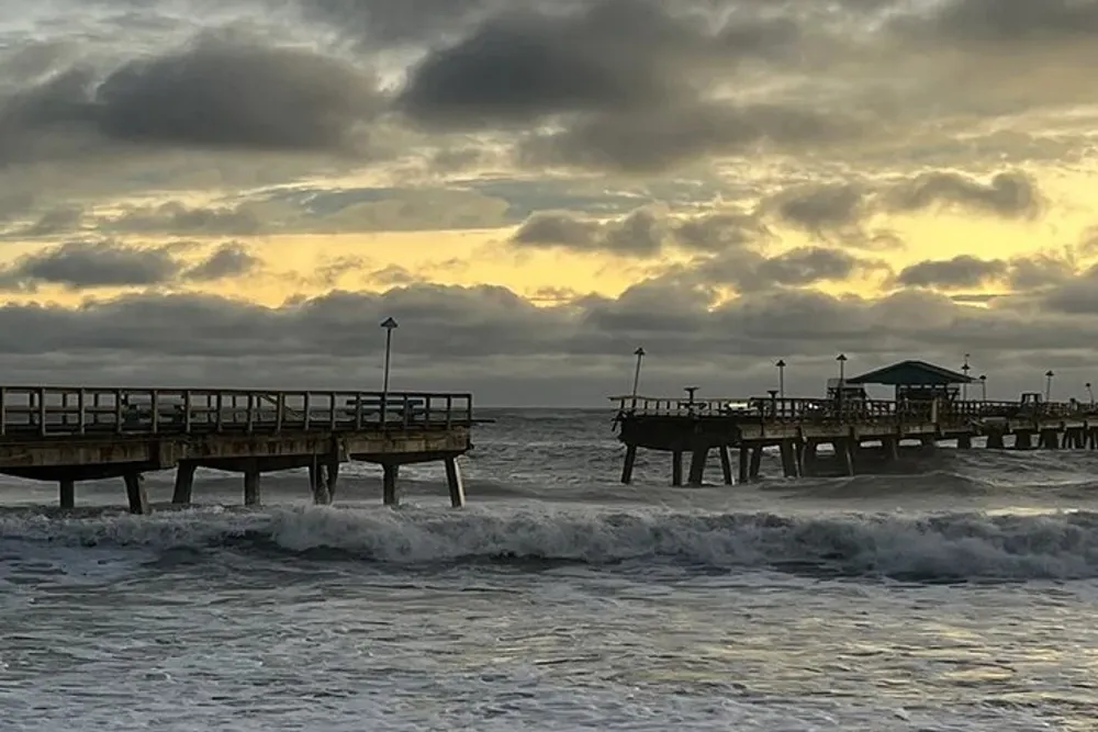 The image features a wooden fishing pier extending into the sea with breaking waves in the foreground and a cloudy sunset sky as the backdrop