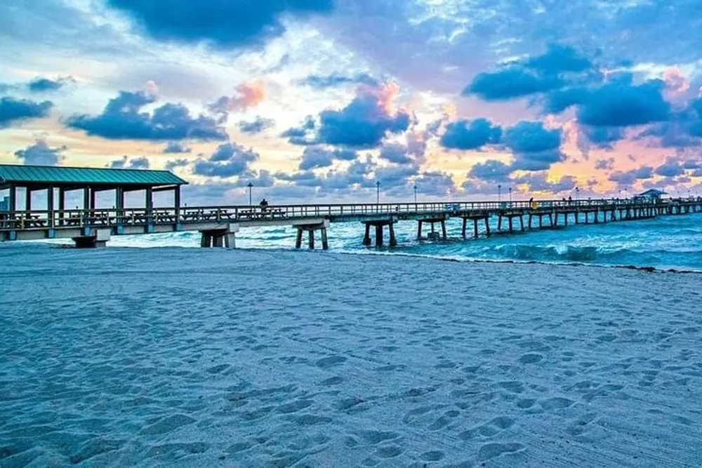 This image depicts a serene beach scene at twilight with a long pier stretching into the sea under a sky painted with pastel-colored clouds