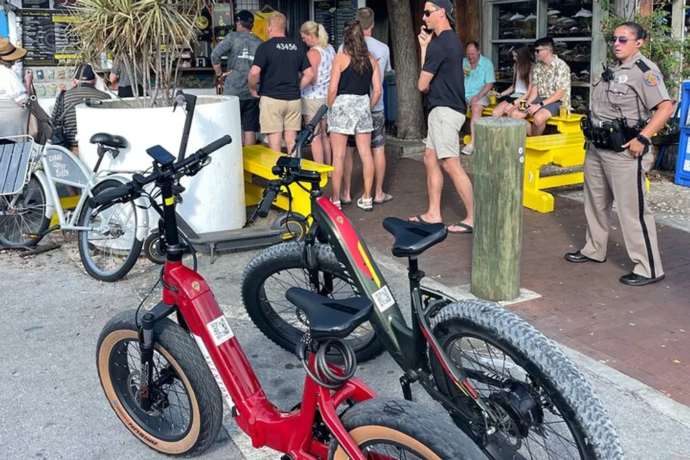 Three electric bikes are parked in the foreground with a group of people and a law enforcement officer standing and conversing in the background likely in a public outdoor casual setting