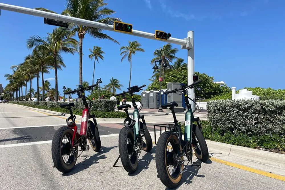 Three electric bicycles are parked by the roadside under a traffic signal on a sunny day with palm trees in the background