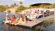 A group of people are enjoying a sunny day on the water, with some sitting on a boat and others on a floating mat near the shore.