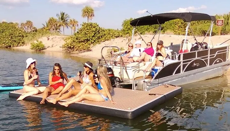 A group of people are enjoying a sunny day on the water, with some sitting on a boat and others on a floating mat near the shore.