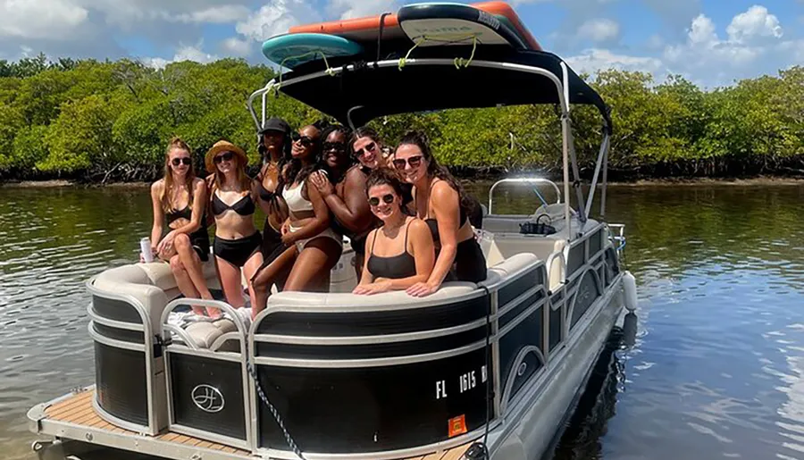 A group of people are enjoying a sunny day on a pontoon boat with a paddleboard attached to the top, smiling for a photo against a backdrop of calm water and mangroves.
