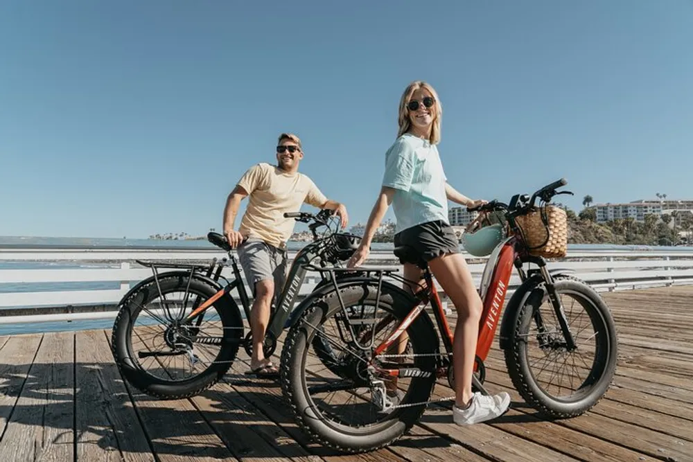 A smiling man and woman stand with bicycles on a sunny boardwalk by the sea