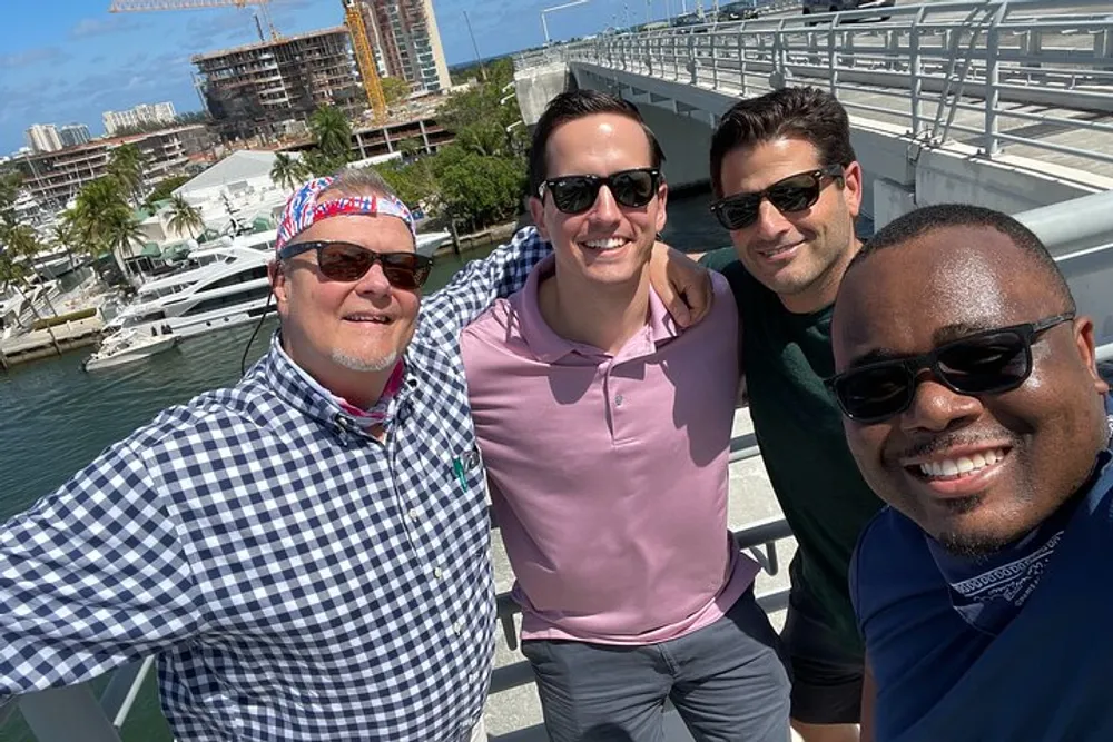 Four smiling men are taking a selfie on a sunny day with a marina and urban construction in the background