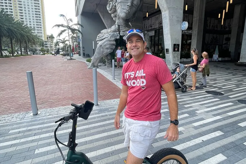 A smiling person in a red t-shirt and white shorts stands next to an electric scooter with a large sculpture and urban surroundings in the background