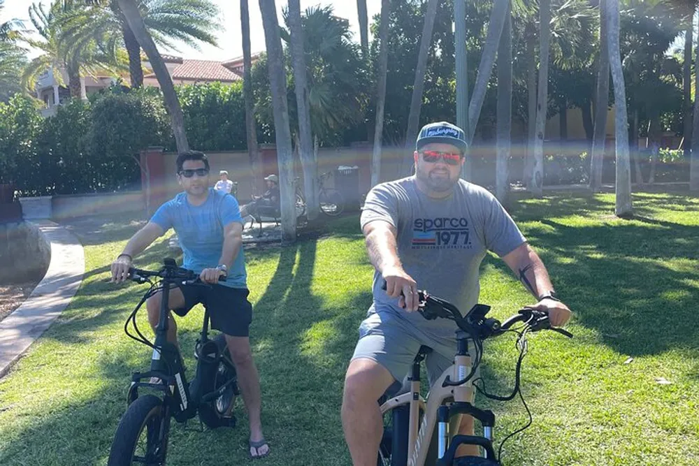 Two people are smiling for the camera while sitting on bicycles in a sunny park with palm trees in the background