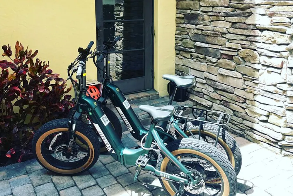 Two electric bicycles with thick tires are parked beside a stone-clad wall near a yellow building