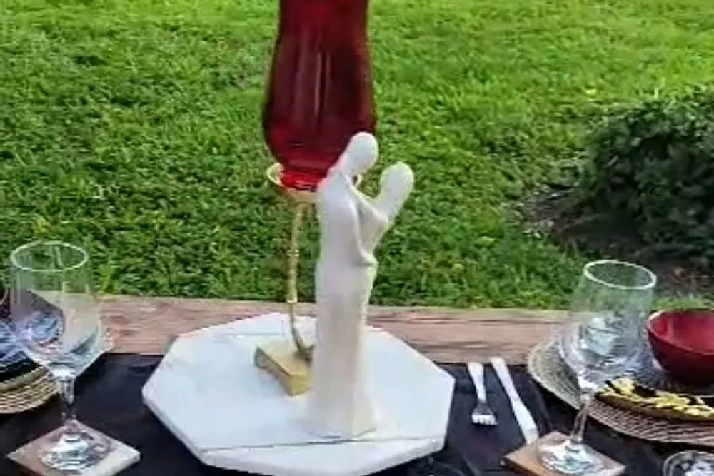 The image shows a creatively designed wine bottle holder shaped like a person with extended arms supporting a tilted bottle of wine set on a table with glasses and snacks in an outdoor setting