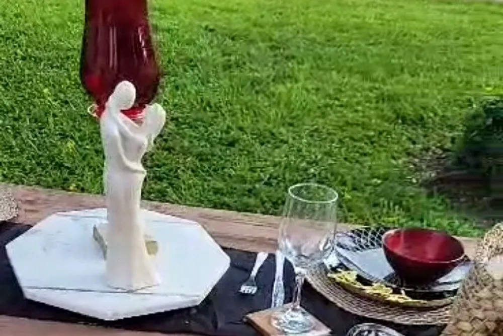 The image shows an elegant outdoor dining setup with a statue of a person a red vase a white plate with utensils a wine glass and a bowl all arranged on a table with a green lawn in the background
