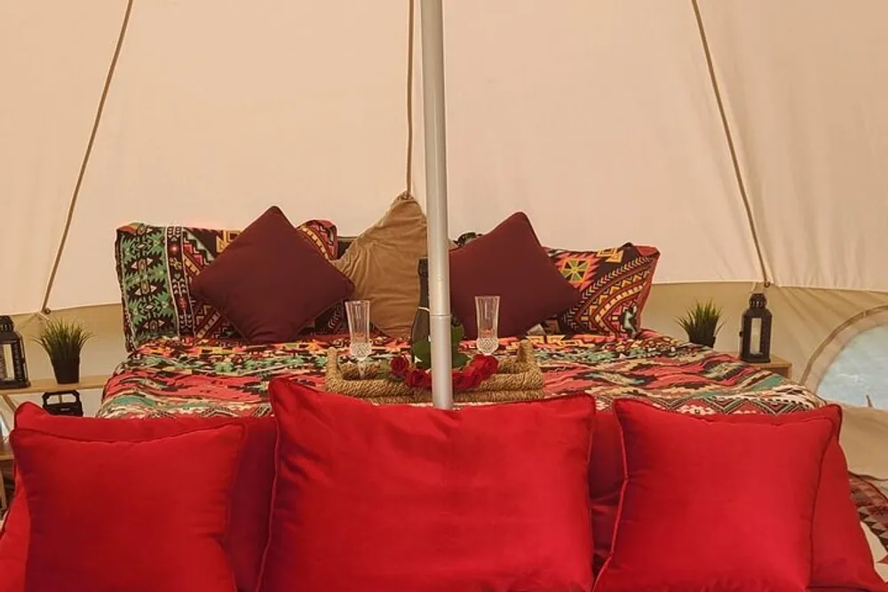 This image depicts a cozy bohemian-style glamping setup inside a tent with vibrant cushions patterned bedding and a romantic table setting with champagne flutes and roses