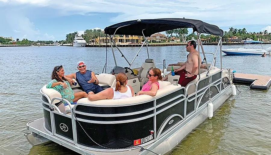 Several people are enjoying a sunny day out on a pontoon boat on calm waters with waterfront homes and another boat visible in the background.