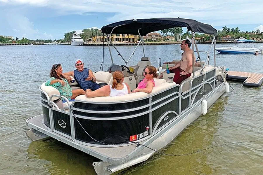 A group of people relax and enjoy a sunny day aboard a pontoon boat on calm waters, with waterfront properties in the background.
