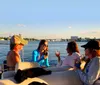 Four people are enjoying a social gathering on a boat with drinks against a backdrop of a calm waterway and city skyline during sunset