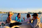 Four people are enjoying a social gathering on a boat with drinks, against a backdrop of a calm waterway and city skyline during sunset.