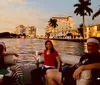 Four people are enjoying a social gathering on a boat with drinks against a backdrop of a calm waterway and city skyline during sunset