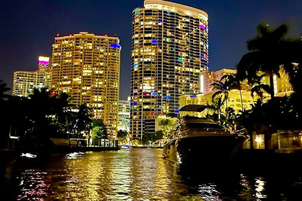 The image depicts a nighttime view of a waterfront with illuminated high-rise buildings a boat docked by the side and the reflections glittering on the surface of the water