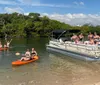A group of people enjoys a sunny day on and around a pontoon boat with some seated aboard the vessel and others on kayaks in shallow waters near a tree-lined shore