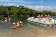 A group of people enjoys a sunny day on and around a pontoon boat, with some seated aboard the vessel and others on kayaks in shallow waters near a tree-lined shore.