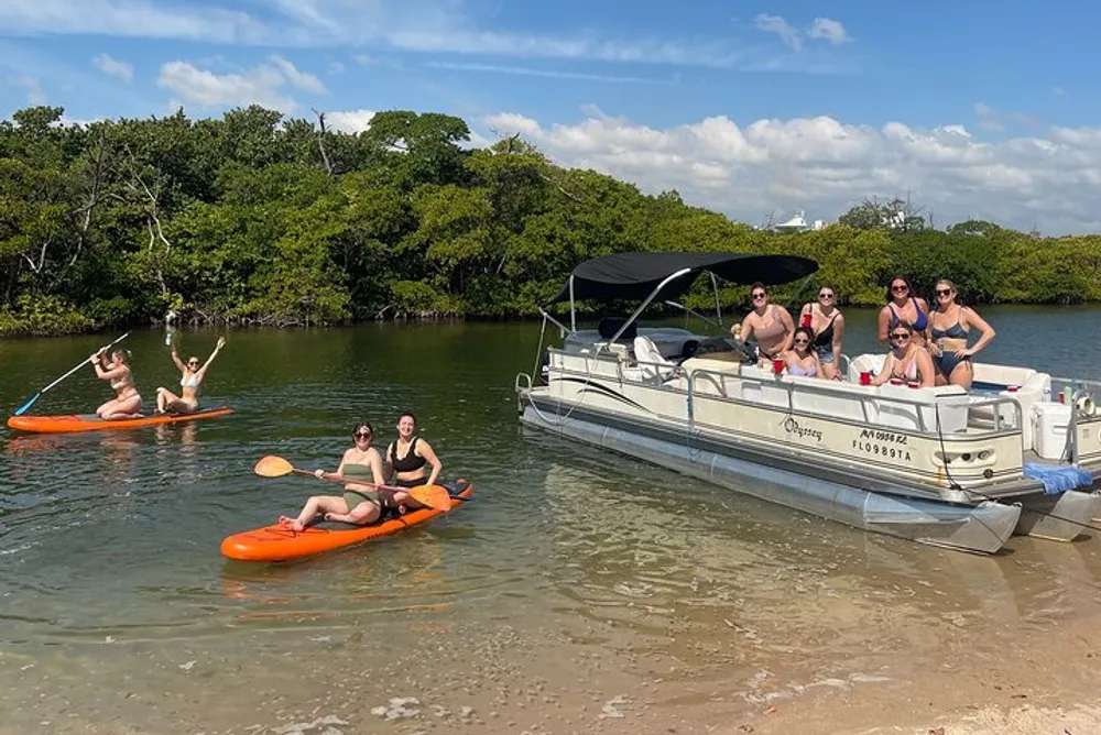 A group of people enjoys a sunny day on and around a pontoon boat with some seated aboard the vessel and others on kayaks in shallow waters near a tree-lined shore