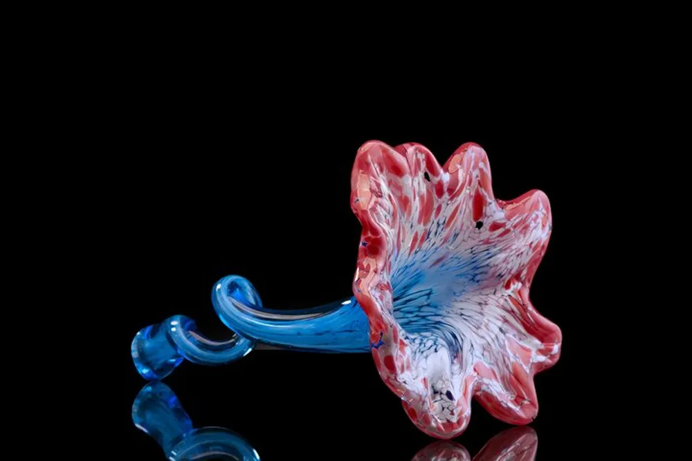 The image shows a striking handcrafted glass sculpture with vibrant red and blue colors set against a reflective black background