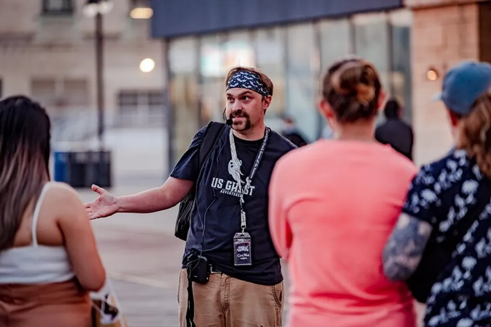 A man wearing a bandana and a lanyard is gesturing as he communicates with a group of people outdoors