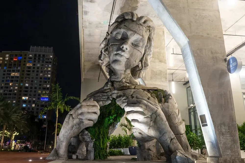 The image shows a large fragmented statue of a womans head and torso partially blended with natural elements like foliage situated under an overpass in an urban environment at night