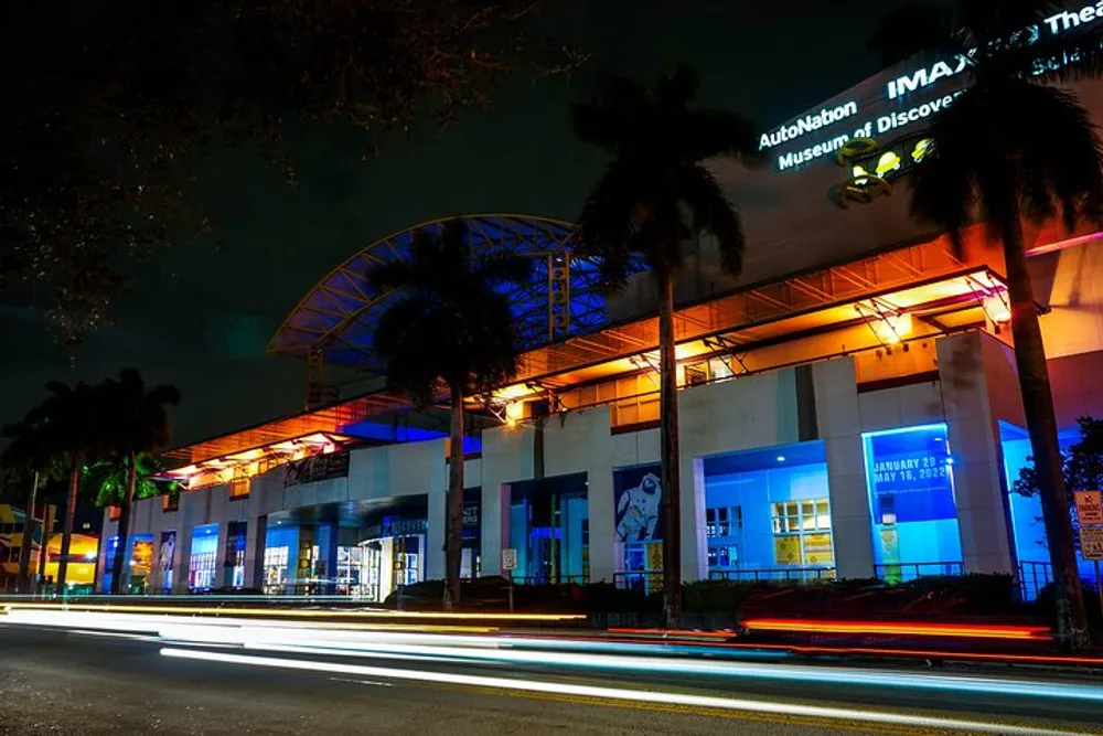 A nighttime view of a museum with vibrant blue lighting at the entrance and light trails from passing traffic in the foreground