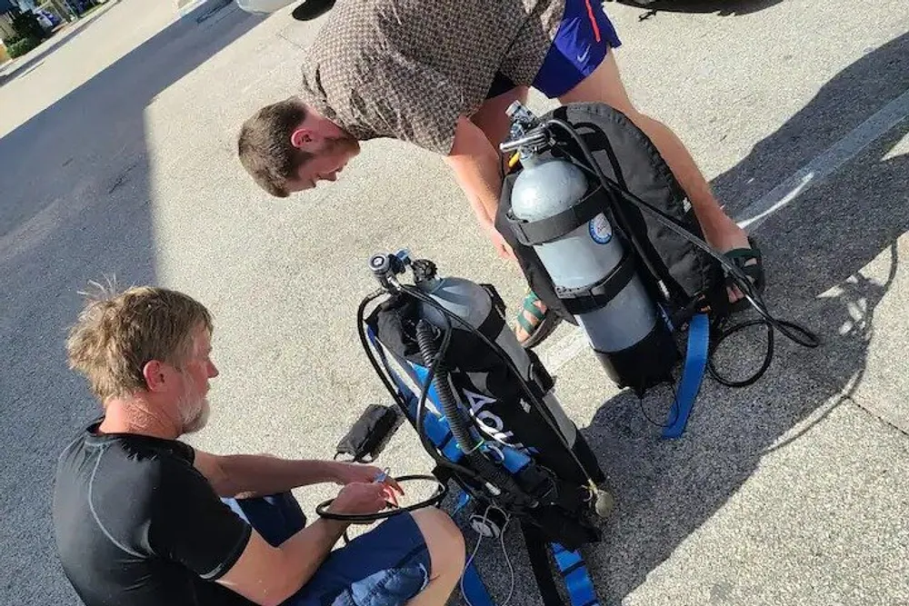 Two individuals are examining or preparing scuba diving equipment on a paved surface