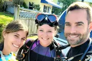 Three people in diving gear are posing for a selfie with bright smiles, suggesting they are about to embark on a scuba diving adventure.