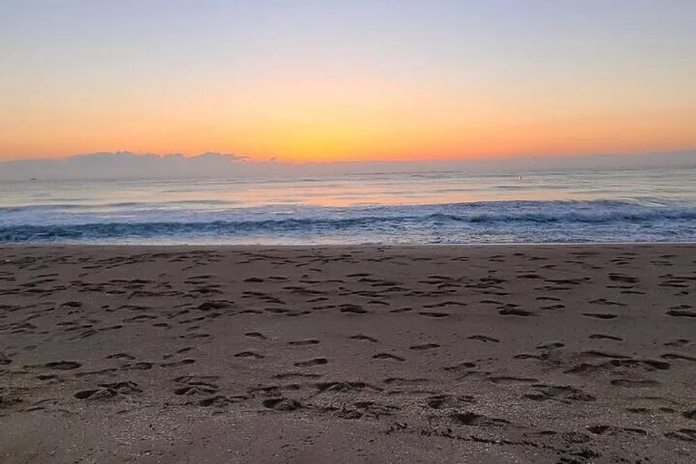The image depicts a serene beach at sunrise with the sky transitioning in hues from blue to orange and the sand imprinted with numerous footprints