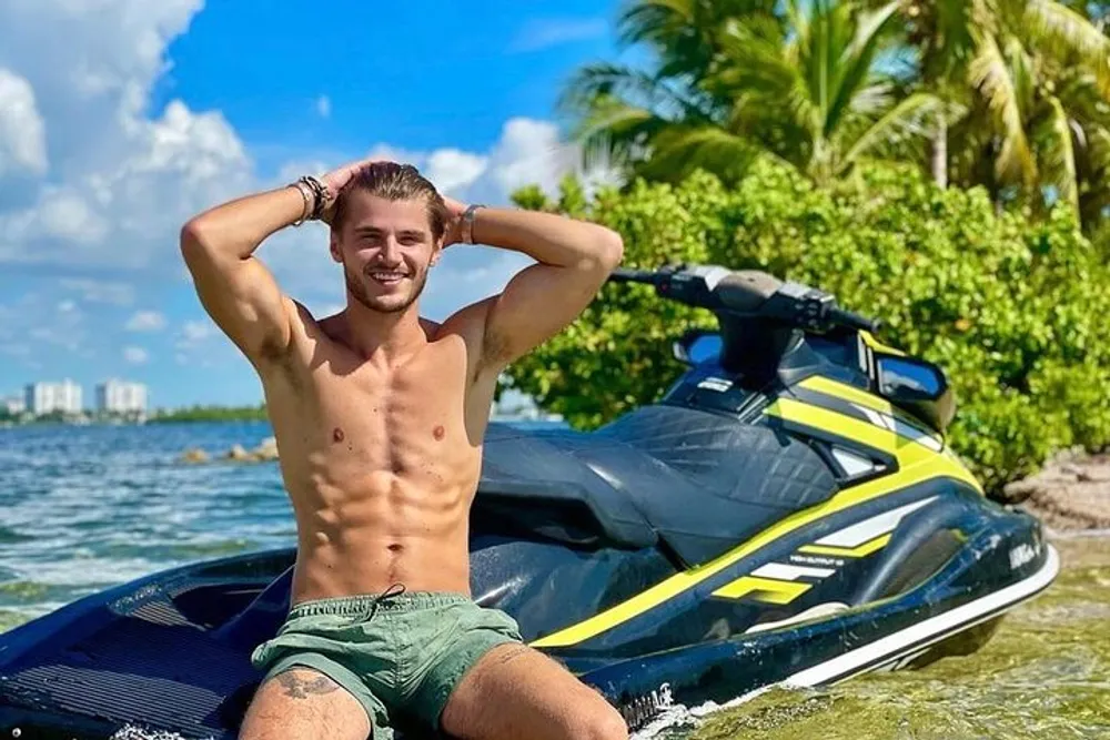 A smiling person is sitting in shallow water near a beach with a jet ski behind him appearing to enjoy a sunny day outdoors