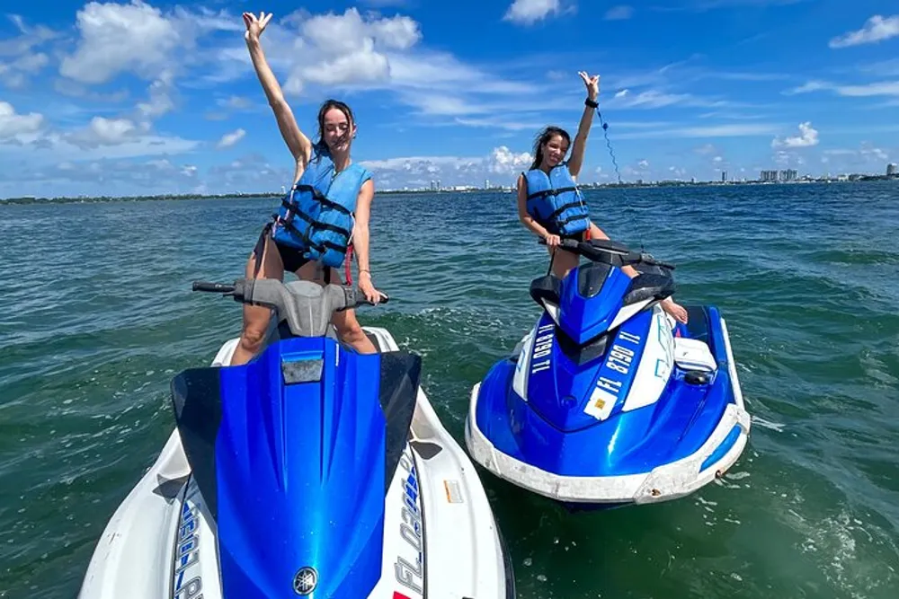 Two people are joyfully posing on jet skis on a sunny day with a city skyline in the background
