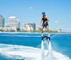 A person is hoverboarding above the water near a coastal cityscape under a clear blue sky