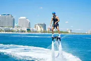 A person is hoverboarding above the water near a coastal cityscape under a clear blue sky.