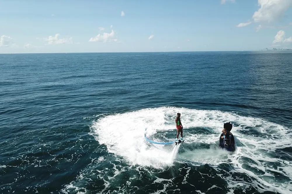 A person appears to be standing on a surfboard while another sits on a jet ski both surrounded by the frothy wake of the jet ski on the open sea
