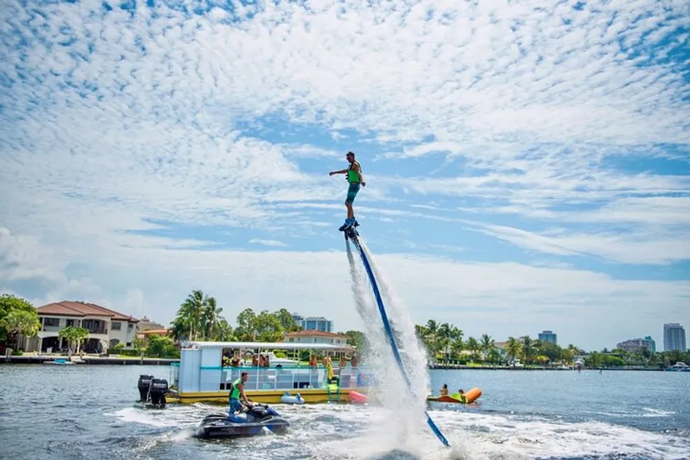 A person is flyboarding high above the water with a jet ski nearby in a sunny scenic waterfront area with scattered clouds in the sky
