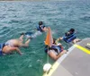 A person is cheerfully raising her arm on a boat while others with snorkeling gear swim in the sea nearby