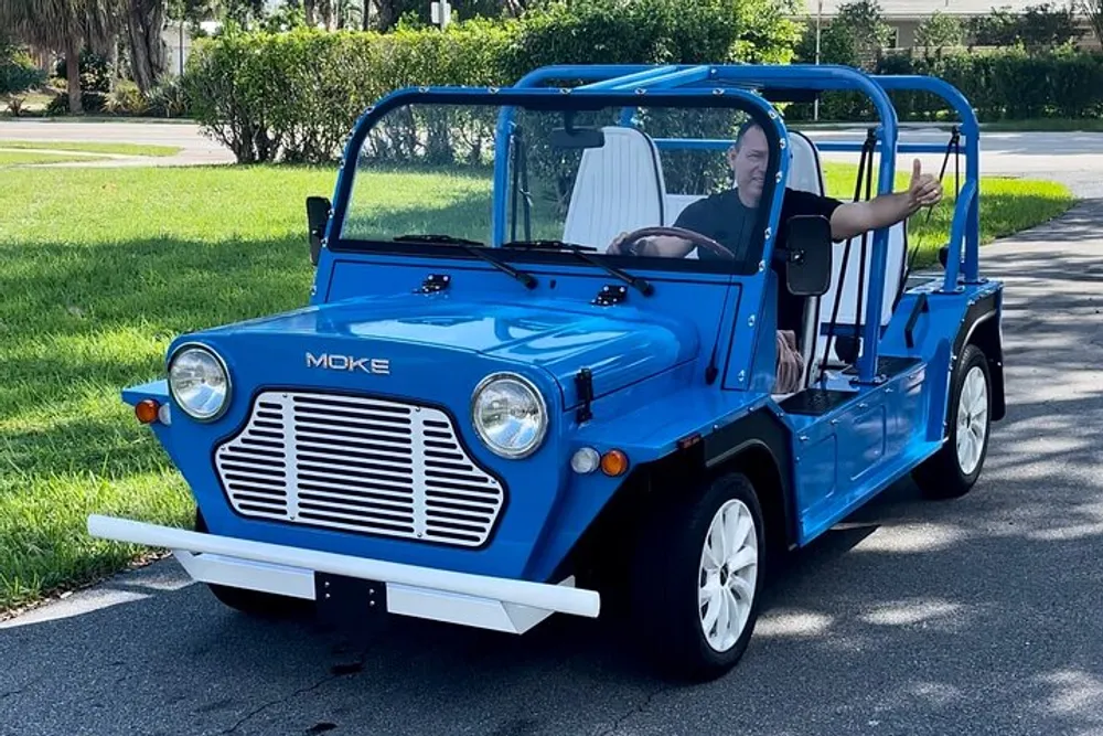 A person is waving from the drivers seat of a bright blue Moke utility vehicle parked on a sunny residential street