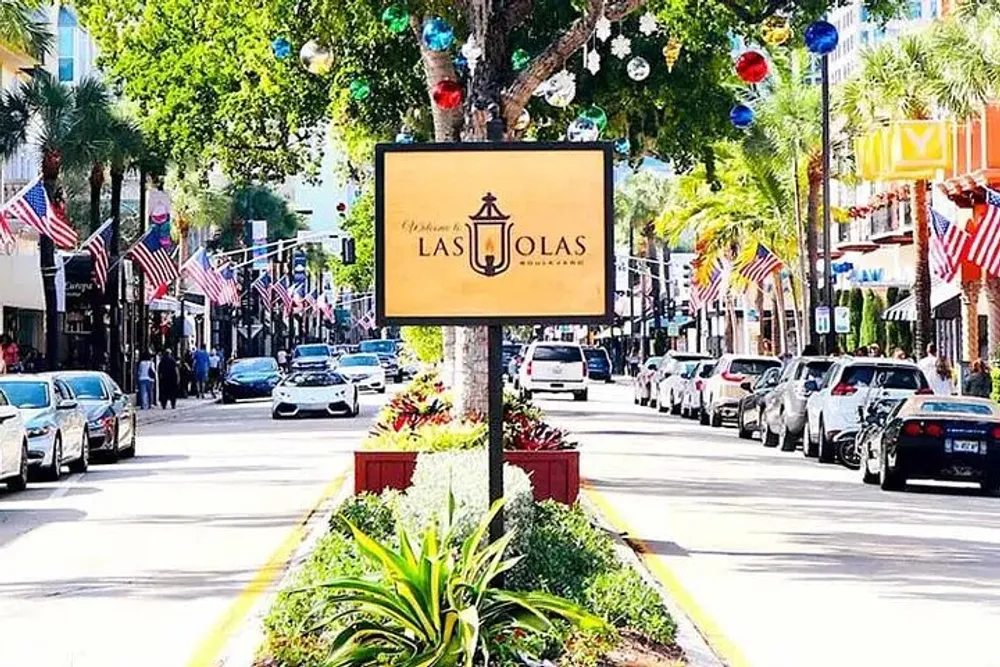 This image shows a sunny street view of Las Olas Boulevard lined with American flags ornamental lighting and parked cars indicating a festive or patriotic atmosphere in what appears to be a shopping or dining district