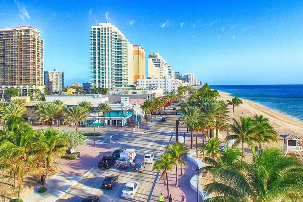 The image shows a sunny coastal road lined with palm trees adjacent to a beach with a backdrop of high-rise buildings under a clear blue sky